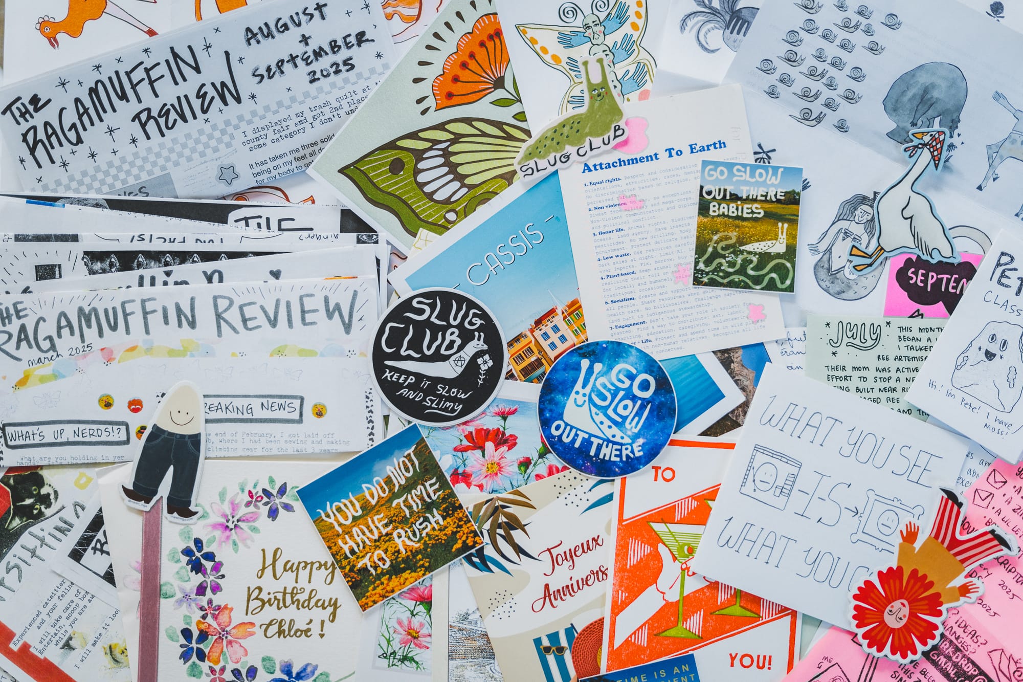 A colorful flat lay of zines, postcards, and stickers, including The Ragamuffin Review and Slug Club, featuring hand-drawn art, bright colors, and handwritten notes.