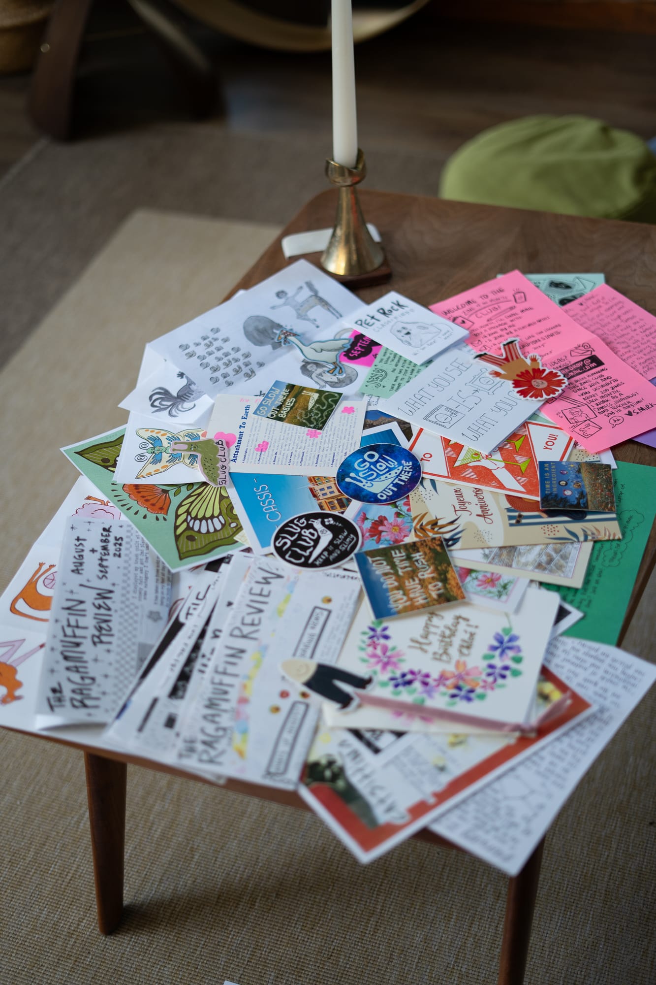 A small wooden table with zines, postcards, and stickers scattered beside a brass candlestick, sunlight highlighting the colorful, hand-drawn mail collection.