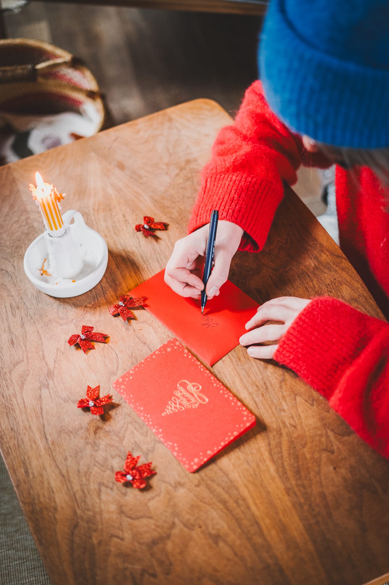 Person in a bright red sweater and blue knit hat writing on a red holiday card at a wooden table, with a lit striped candle in a white holder and small red glitter bows scattered nearby.