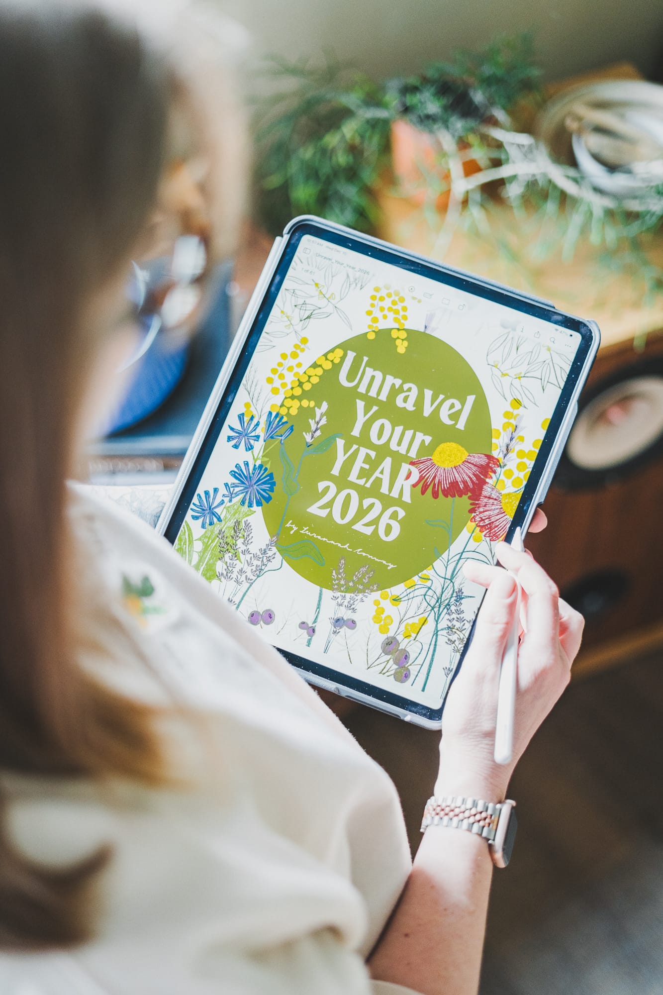A person holds an iPad displaying the illustrated cover of Unravel Your Year 2026 by Susannah Conway, using a stylus while sitting indoors with soft light and plants in the background.