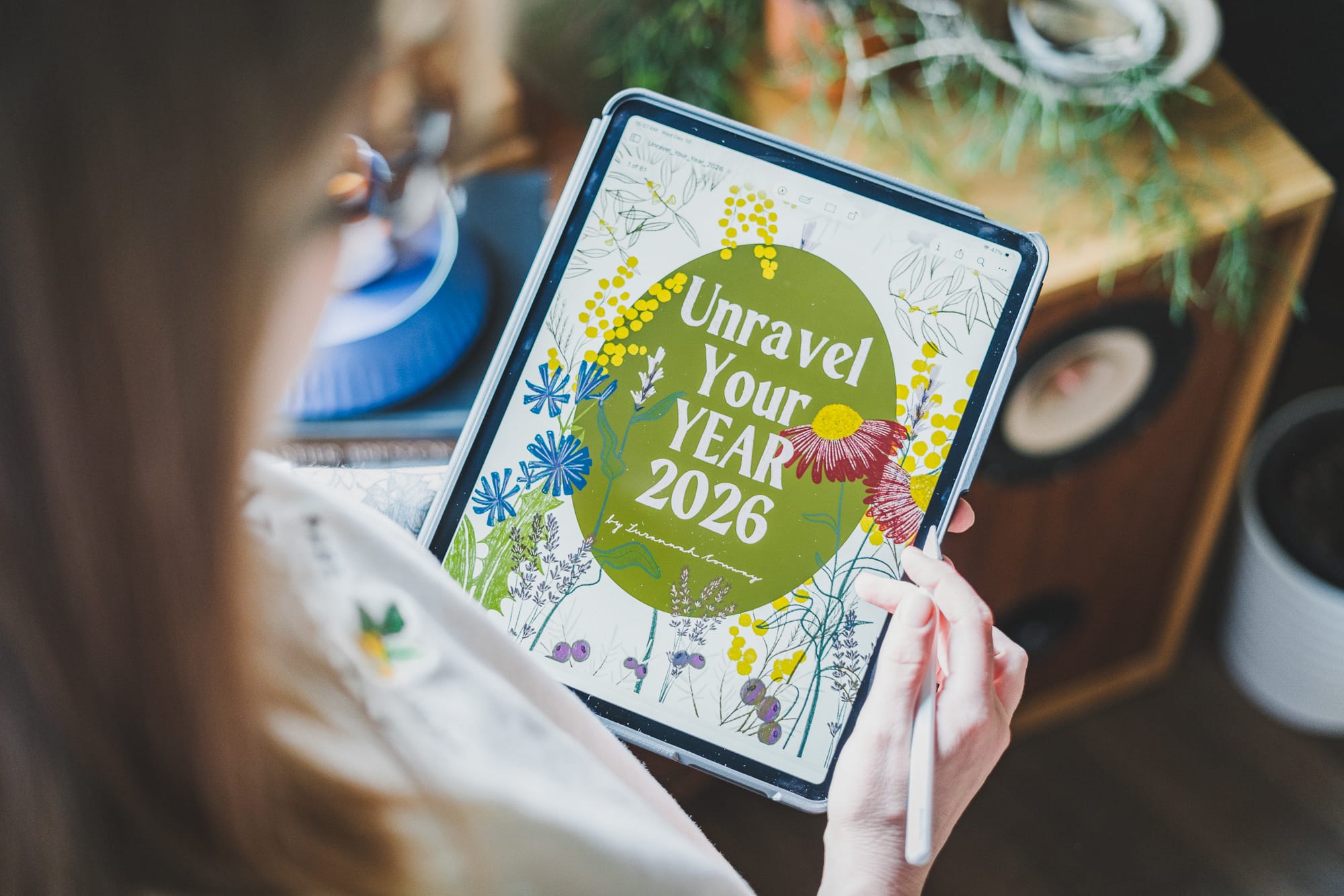 A person holds an iPad displaying the illustrated cover of Unravel Your Year 2026 by Susannah Conway, using a stylus while sitting indoors with soft light and plants in the background.