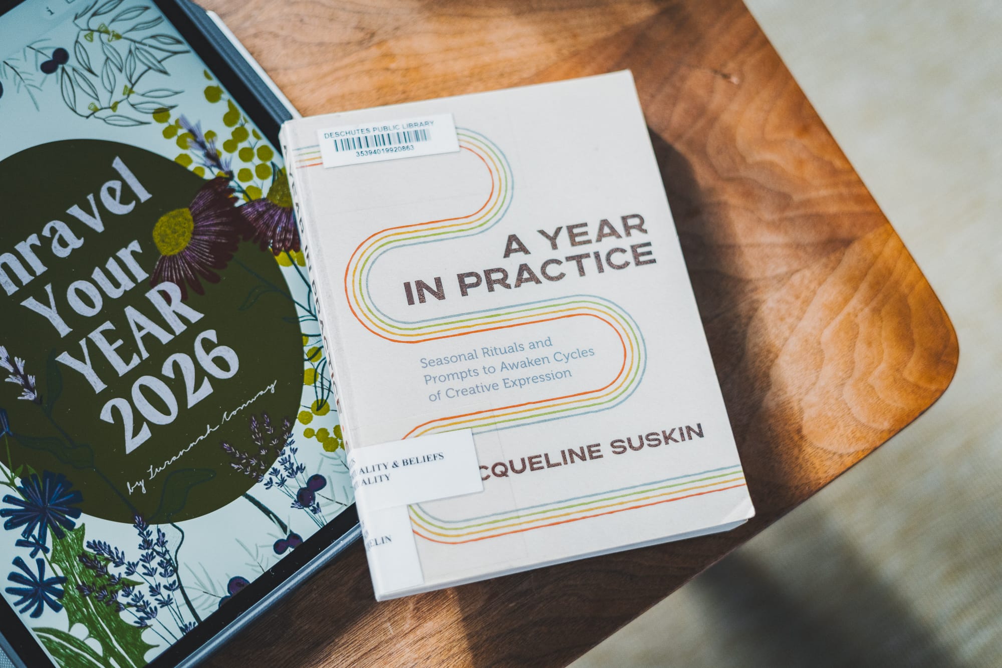 A Year in Practice by Jacqueline Suskin rests on a wooden table beside the Unravel Your Year 2026 workbook, both lit by soft natural light.