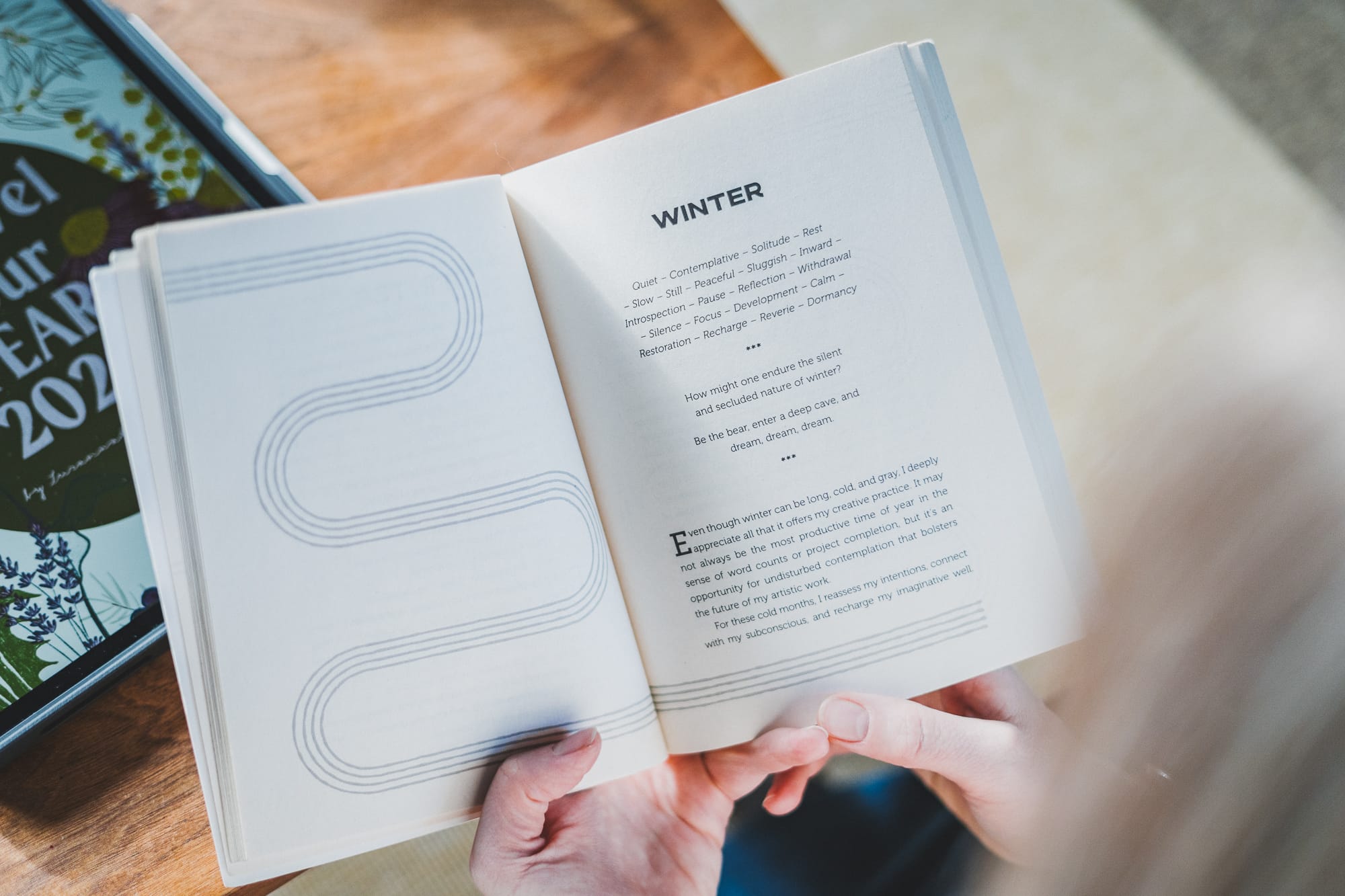 Hands hold A Year in Practice open to the “Winter” chapter, showing seasonal themes like rest, introspection, and calm, with the workbook visible nearby on the table.
