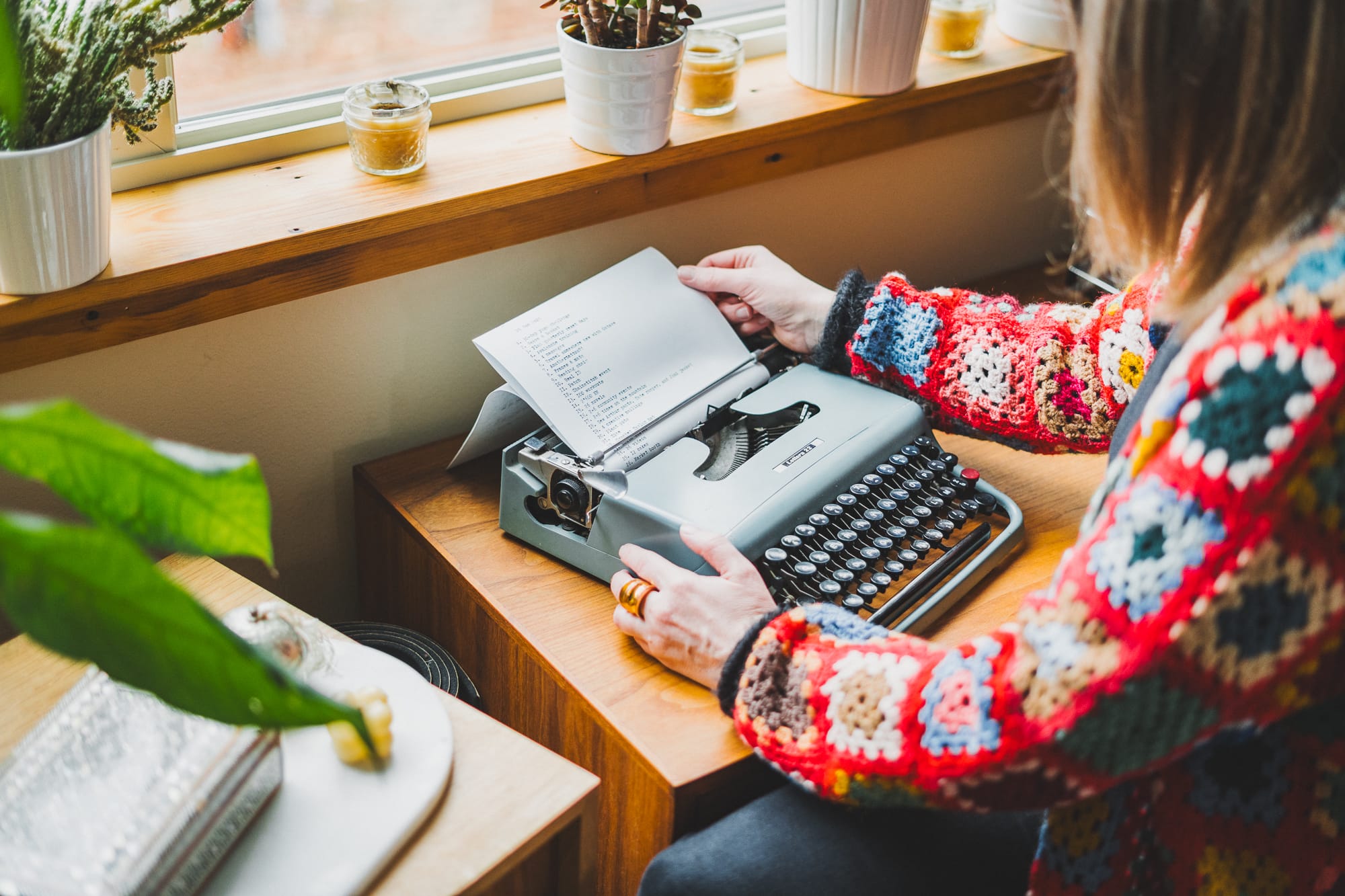 Person wearing a colorful crocheted sweater types a list on a vintage typewriter by a window, with small plants and candles on a wooden windowsill.