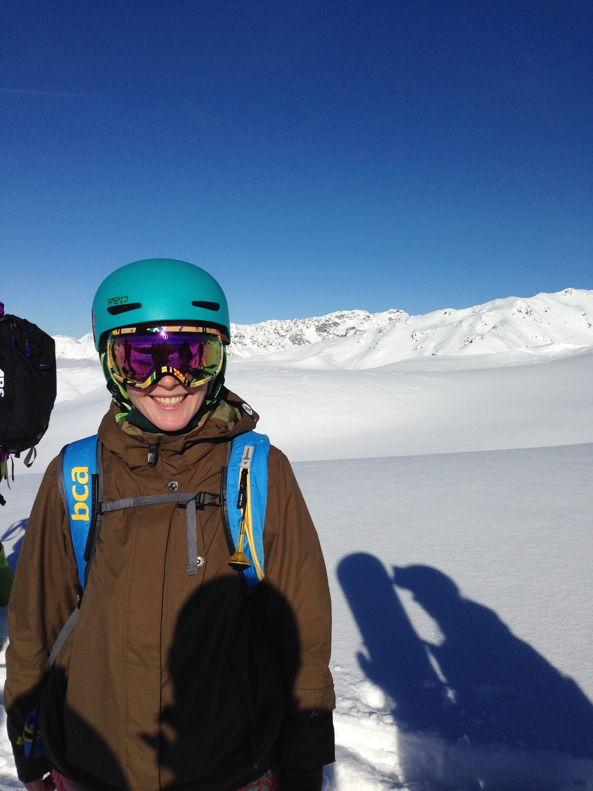 Smiling splitboarder wearing a helmet and goggles stands in an open alpine bowl with snow-covered mountains in the background.