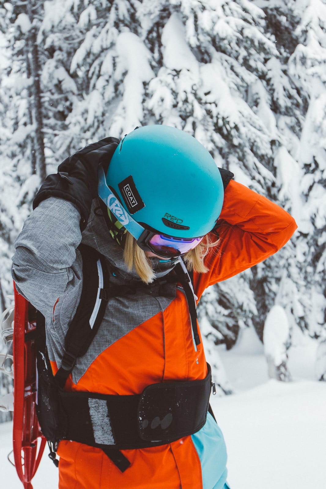 Splitboarder wearing a teal helmet and orange jacket adjusts her pack straps in a snowy forest during a backcountry tour.
