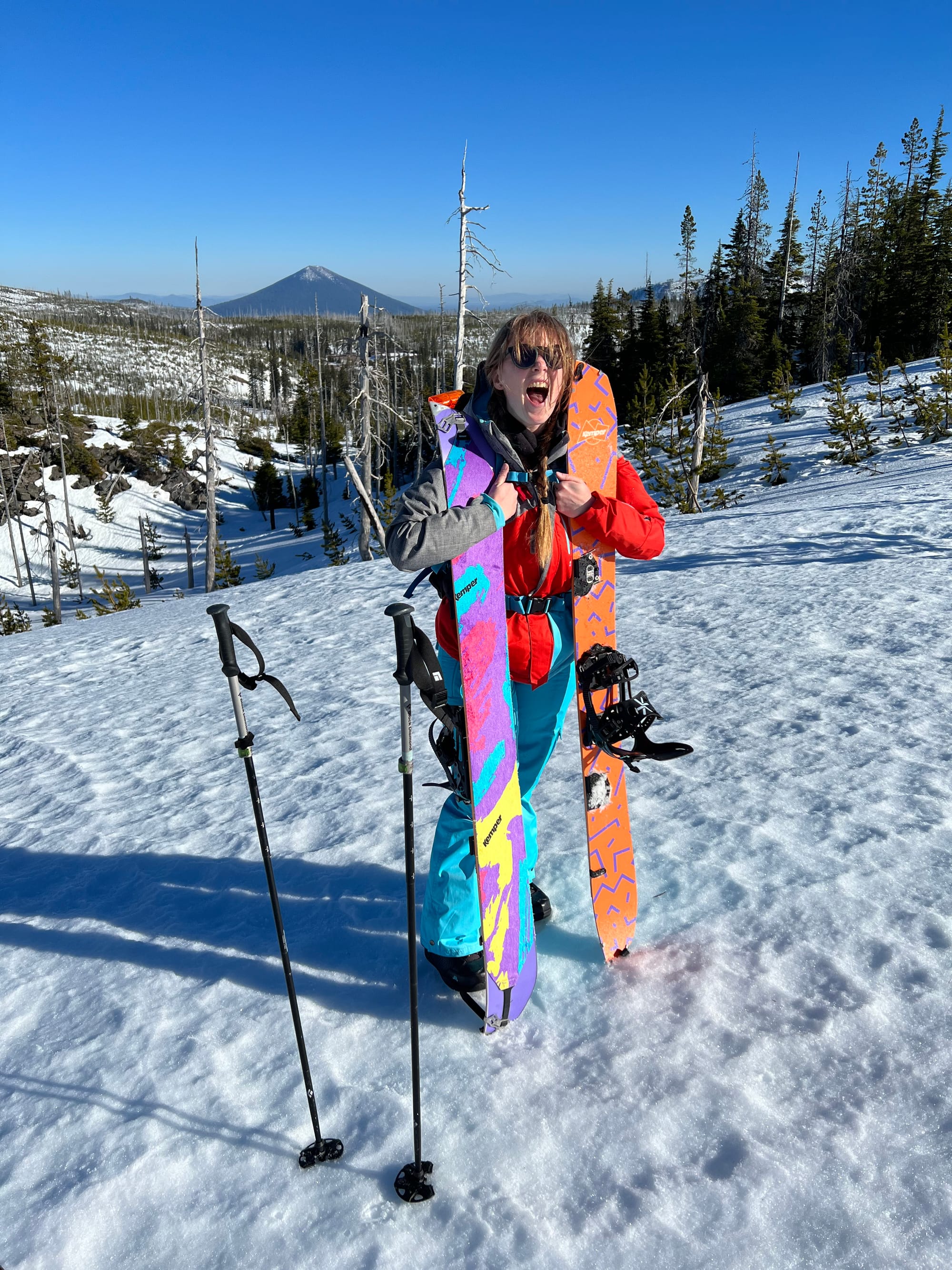 Splitboarder stands holding splitboard skis upright during a break on a sunny backcountry tour, forest and peaks behind.