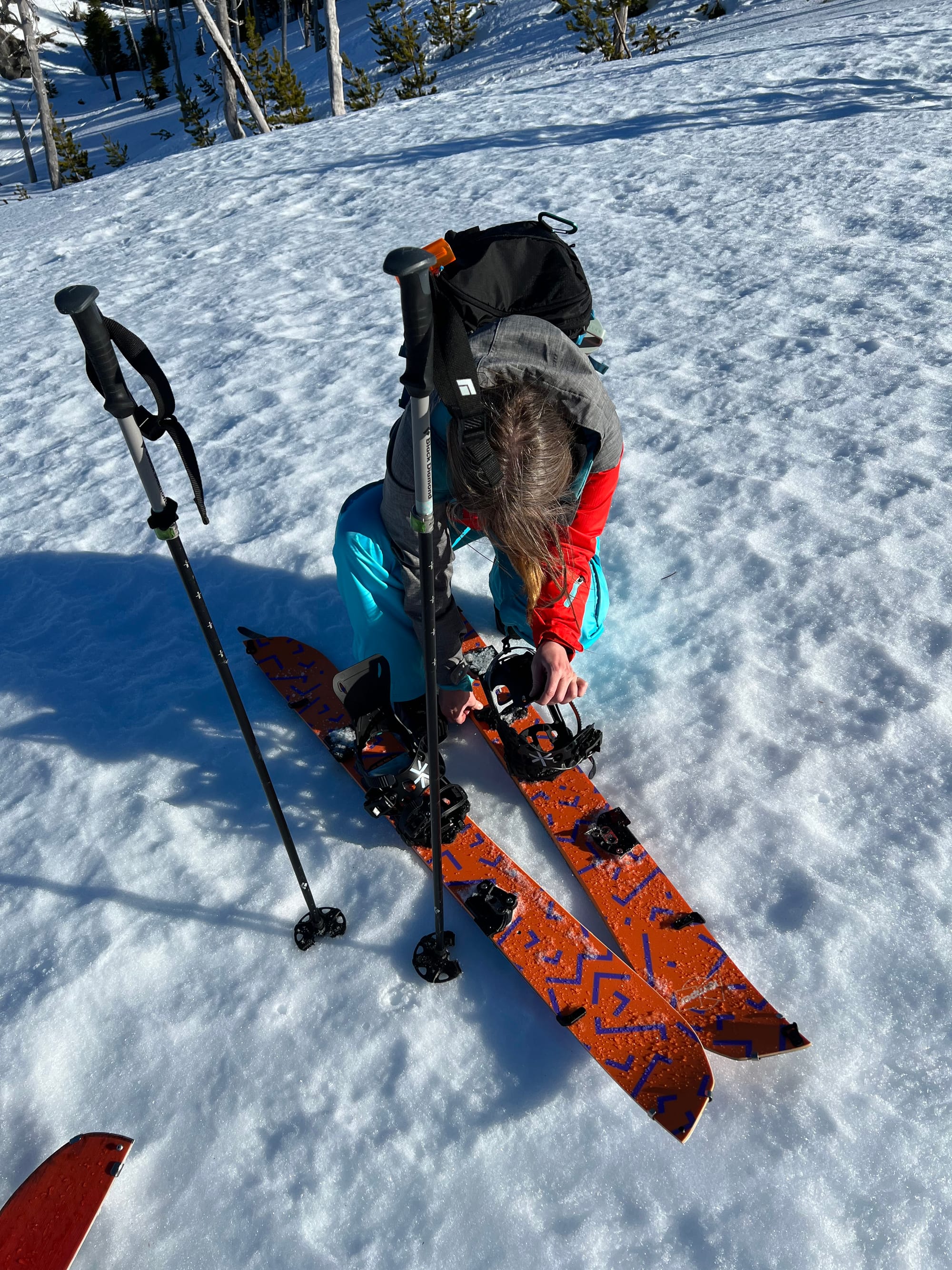 Splitboarder kneels in the snow to adjust bindings while skis are separated in touring mode, with poles planted beside her.