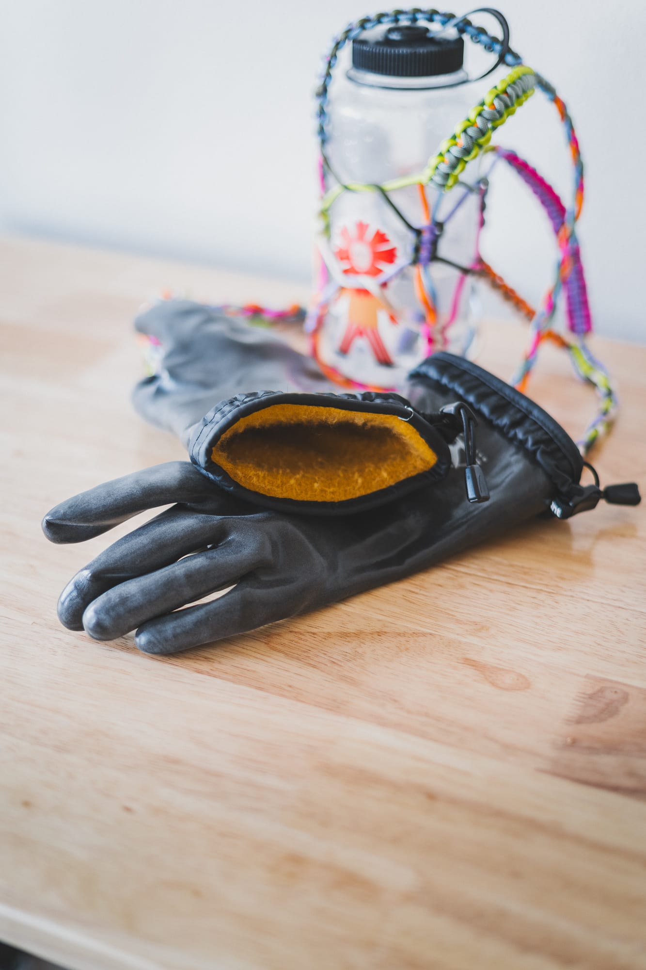 Close-up of black insulated gloves with a mustard fleece lining resting on a wooden table, with a clear water bottle wrapped in colorful braided cord in the background.