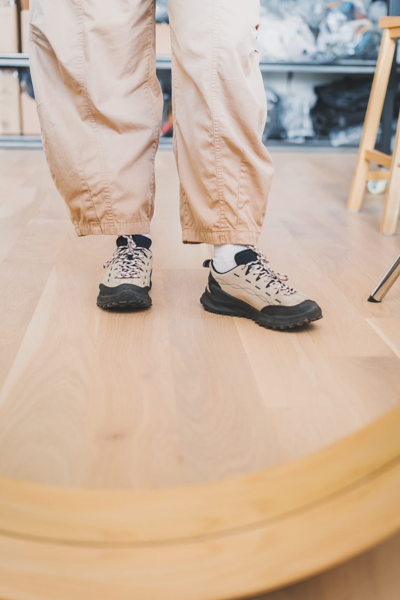 Close-up of beige-and-black hiking sneakers worn with black-and-white socks and tan pants, standing on a light wood floor indoors.