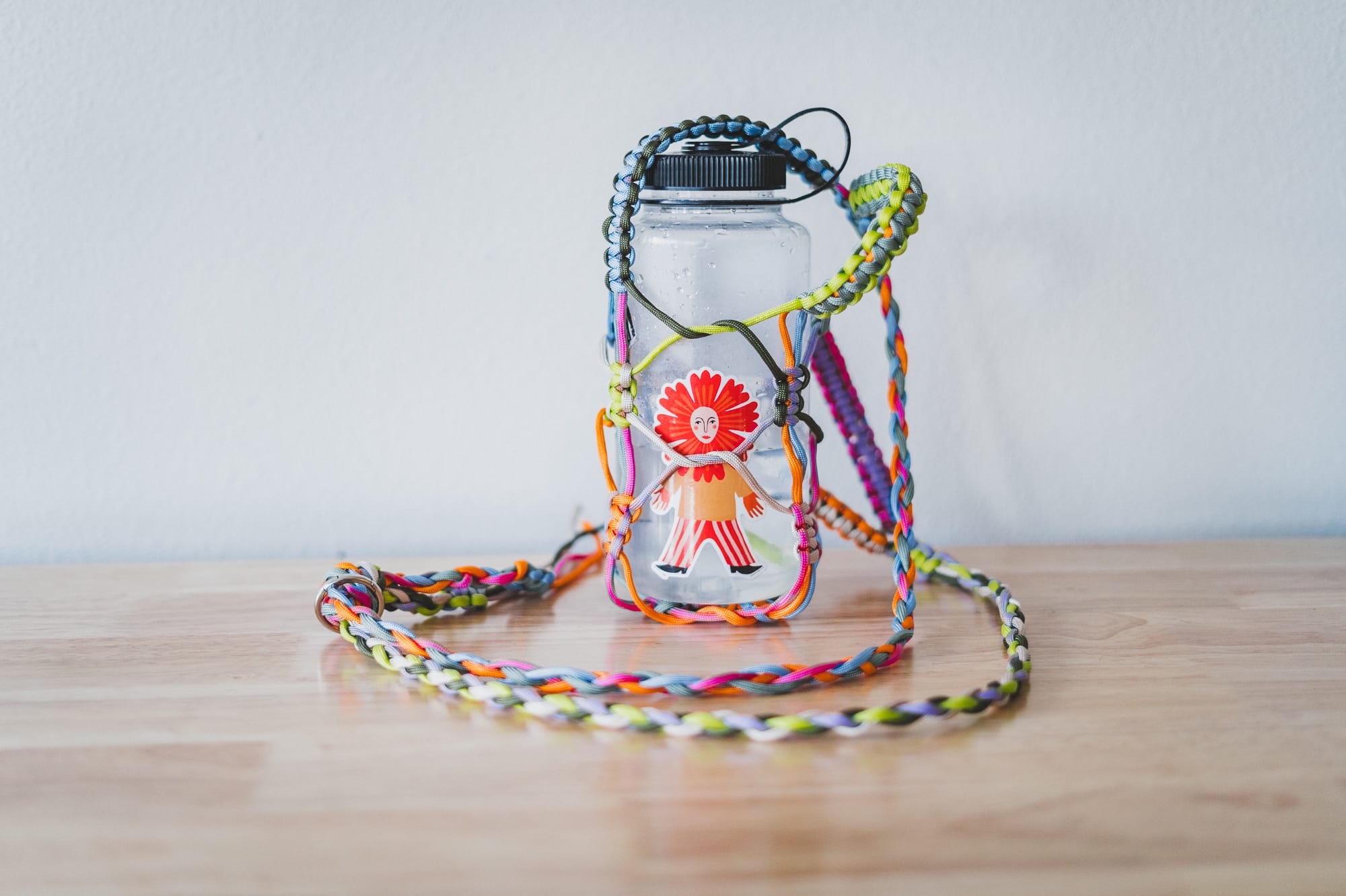 Clear water bottle with a black lid wrapped in a colorful braided paracord holder and long strap, resting on a wooden table against a plain wall.