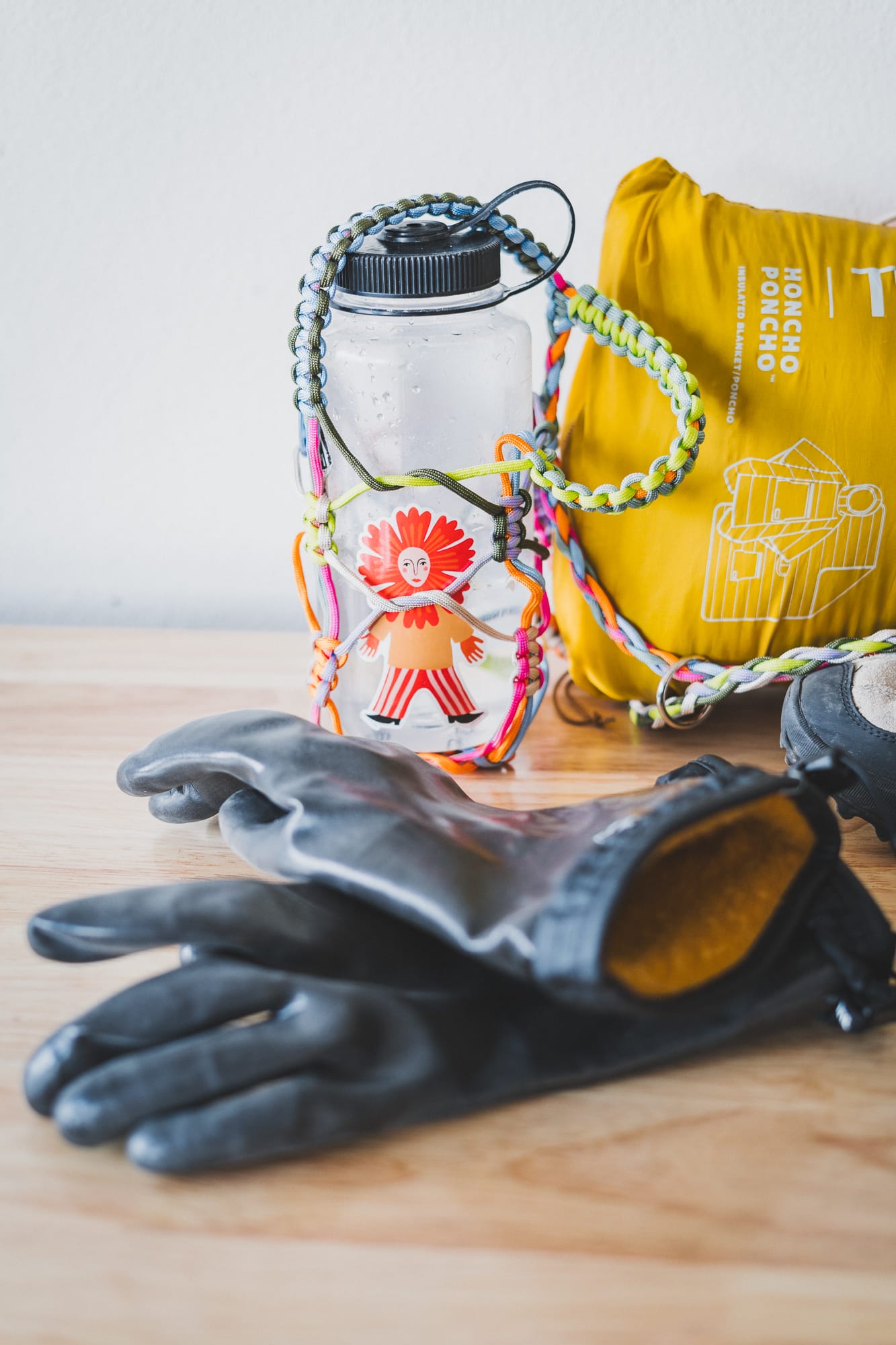 Black insulated gloves with mustard lining rest on a wooden table beside a clear water bottle in a colorful braided paracord holder and a bright yellow packed poncho.