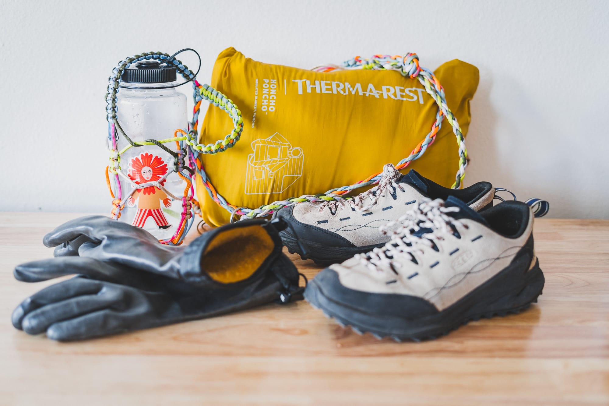 Winter gear on a wooden table: beige-and-black hiking shoes, black insulated gloves, a clear water bottle with colorful braided paracord strap and sticker, and a yellow  poncho.