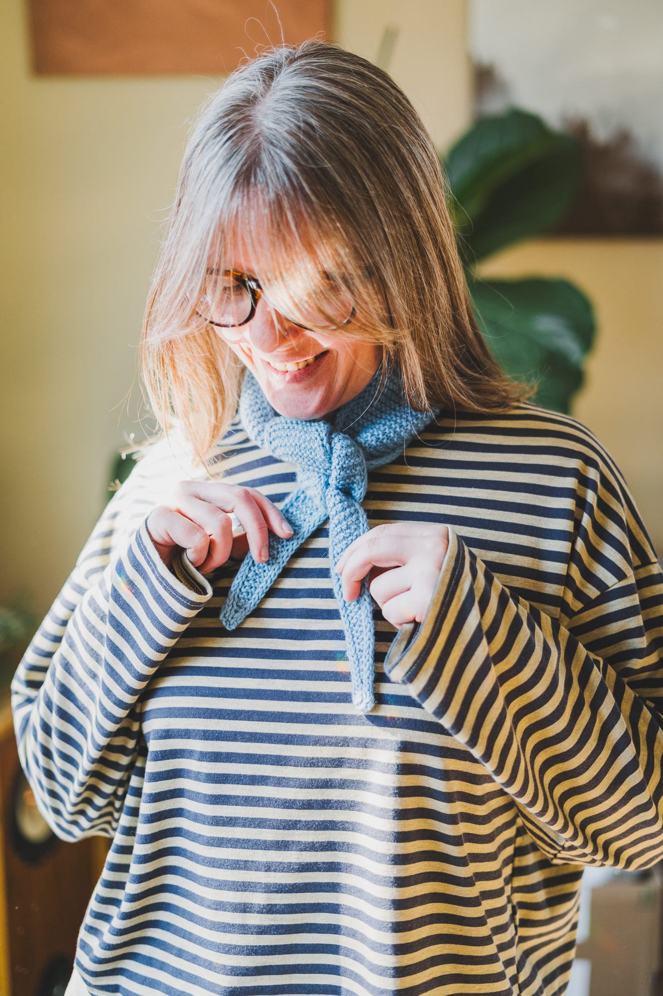 Woman wearing a light blue hand-knitted Sophie Scarf by PetiteKnit tied at the neck, styled over a striped shirt and shown as a finished knitting project.